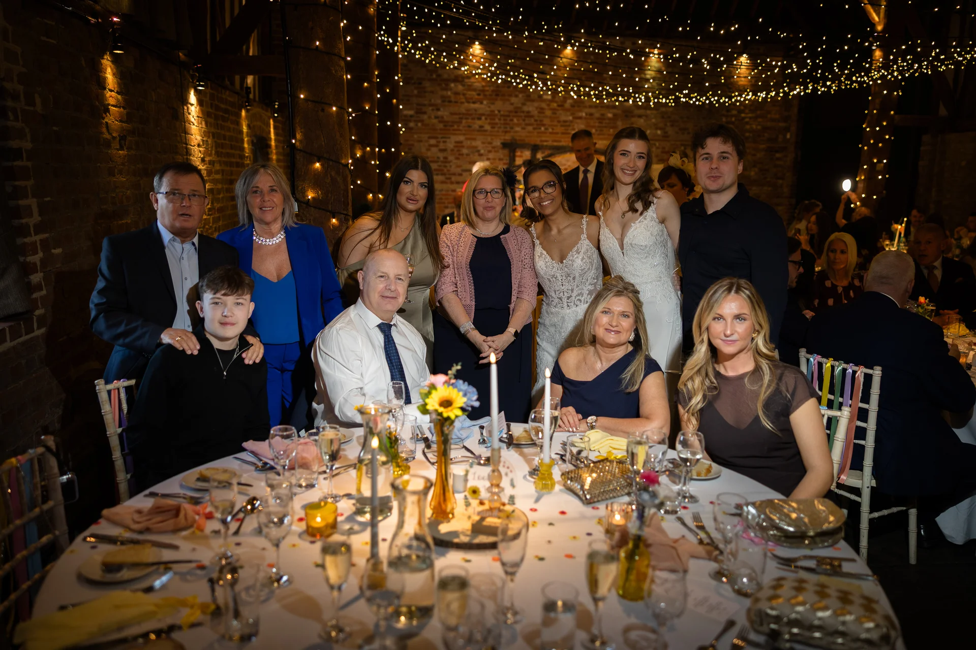 A large group of wedding guests and family smile together around a candlelit round table inside a rustic brick barn adorned with a twinkling fairy light canopy.