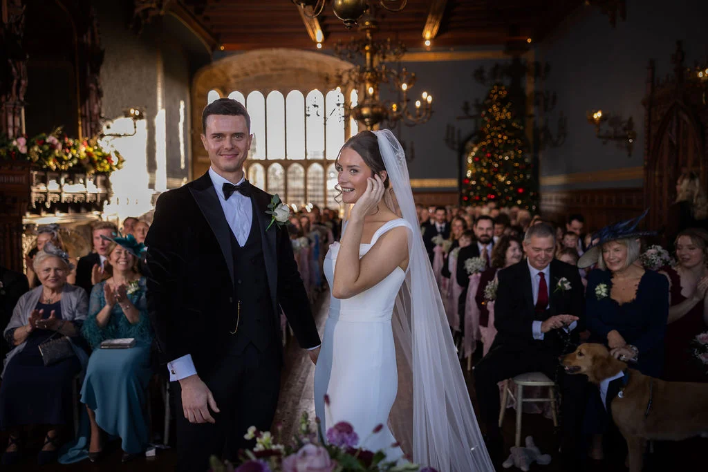 Newlyweds stand smiling at the altar in a grand wood-panelled hall filled with seated guests and a glowing Christmas tree.