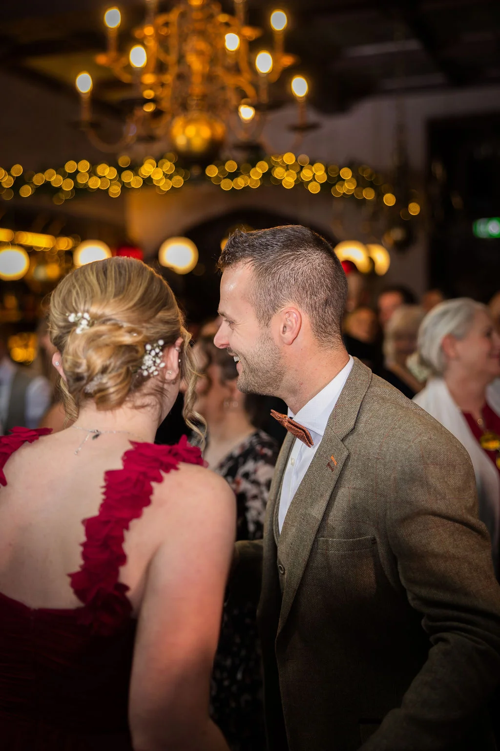A smiling groom in a tweed jacket and bow tie dances with his bride, who wears a striking red dress featuring ruffled straps, beneath a glowing chandelier and fairy lights in a pub reception venue.