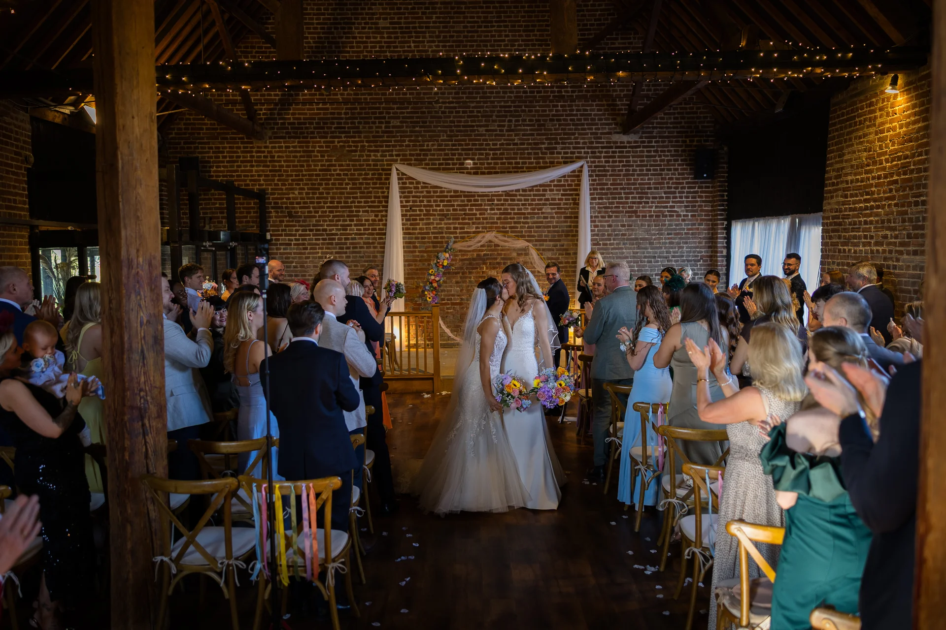 Two brides share their first kiss as a married couple at the end of the aisle in a rustic brick barn filled with applauding guests.