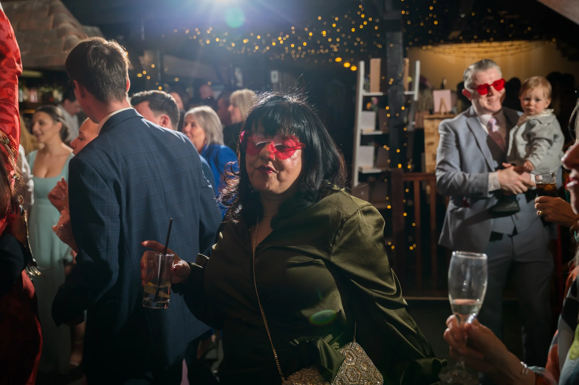 A guest wearing heart-shaped sunglasses enjoys a drink while mingling with others during the lively evening wedding reception.