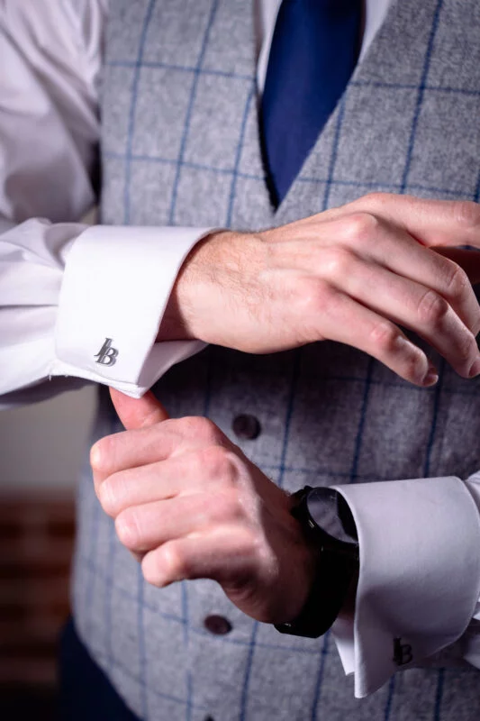 A groom carefully adjusts his monogrammed silver cufflinks while wearing a grey checked tweed waistcoat and a blue tie on the morning of his wedding.