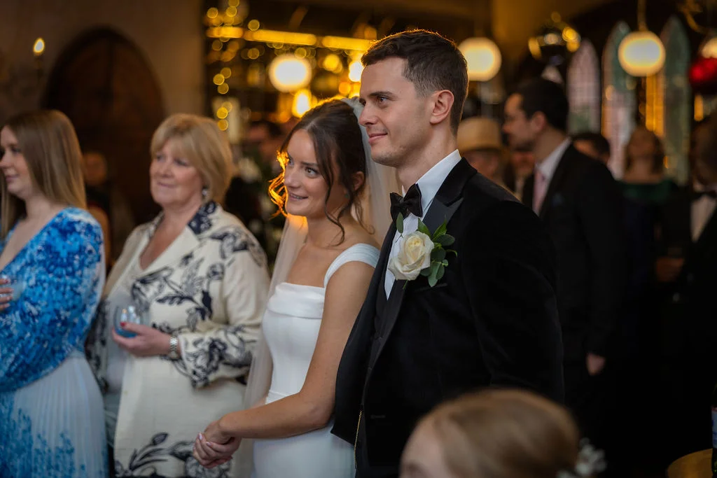 A smiling bride and groom stand together in a warm, candlelit indoor venue surrounded by wedding guests during the ceremony.