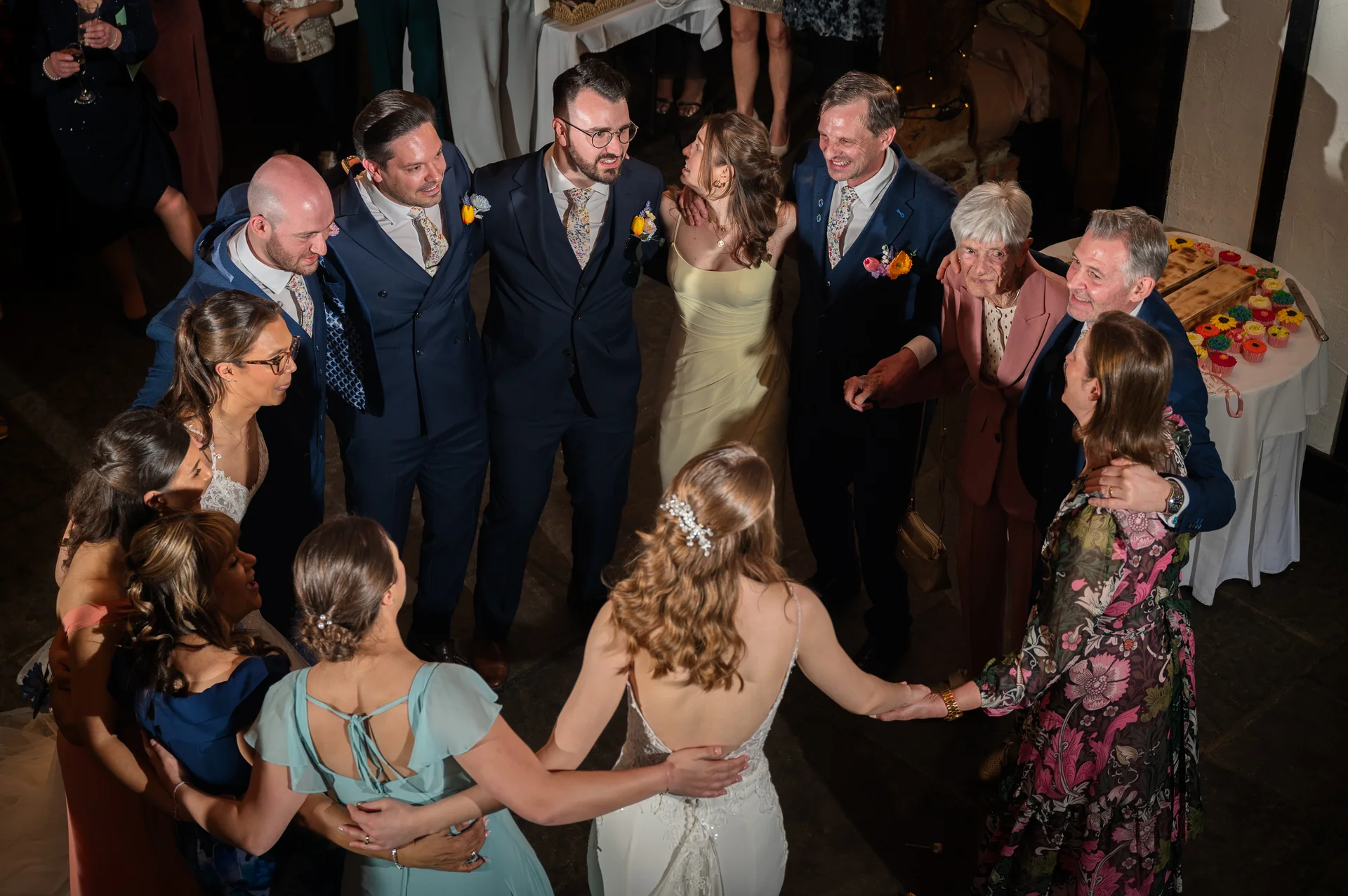 A high-angle candid shot of a tightly knit circle of wedding guests and family members linking arms and laughing joyfully together at a reception.