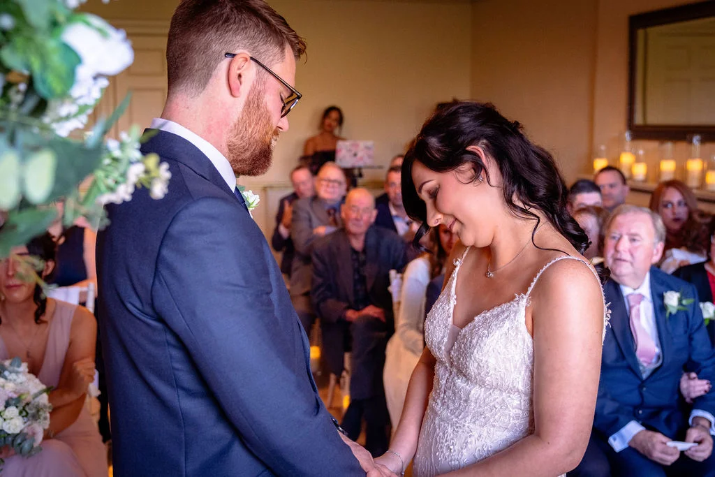 The bride and groom stand facing each other during their emotional wedding ceremony, holding hands while surrounded by seated guests.