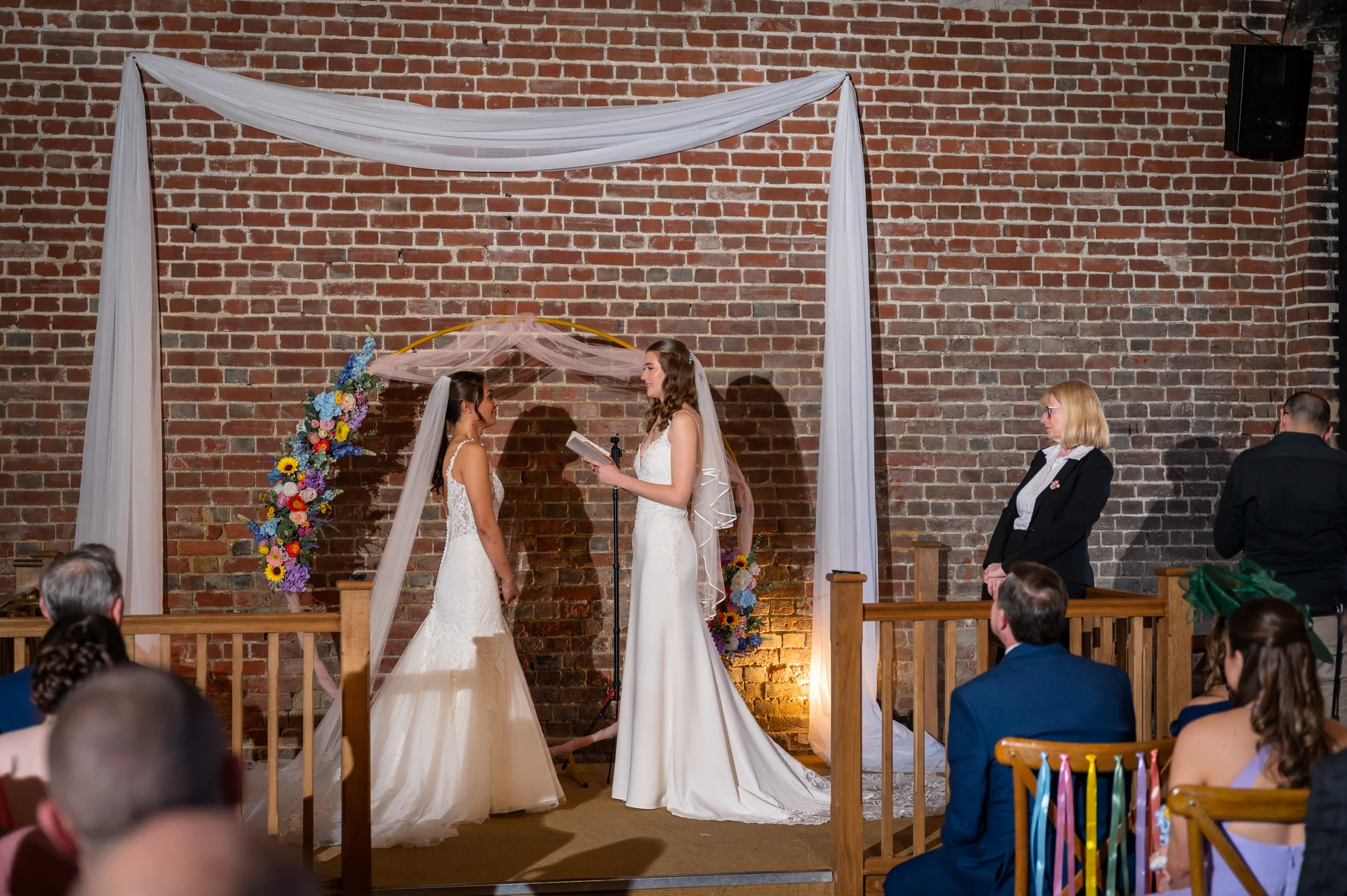 Two brides stand facing each other to read their vows during an indoor wedding ceremony against an exposed brick wall adorned with white draping and floral arrangements.