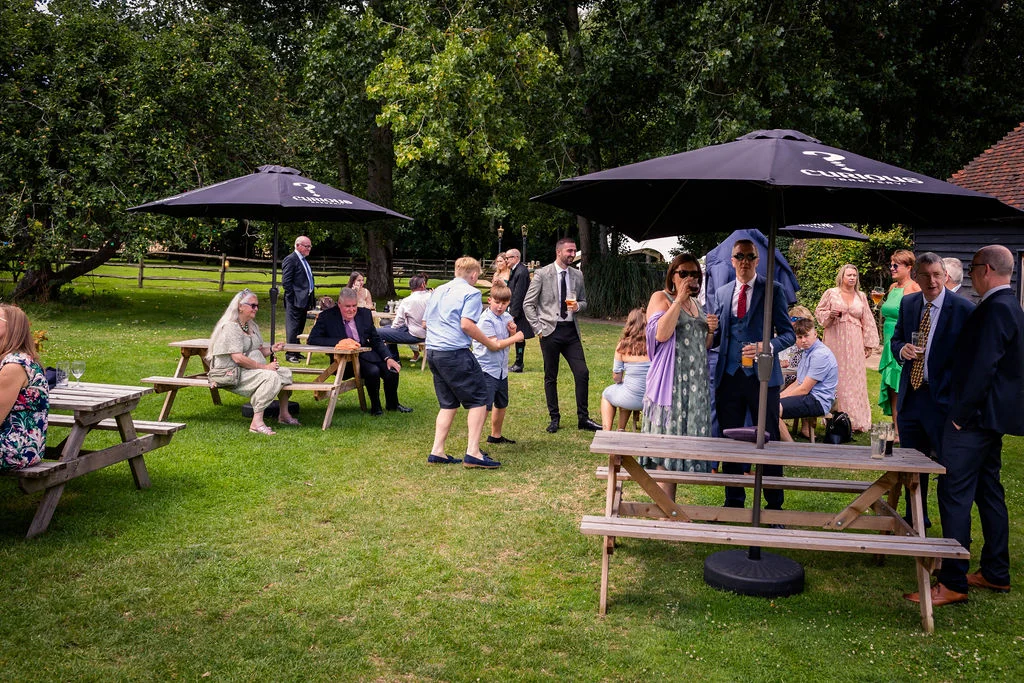 Wedding guests mingle and relax on the lawn outside a venue during an outdoor summer drinks reception.