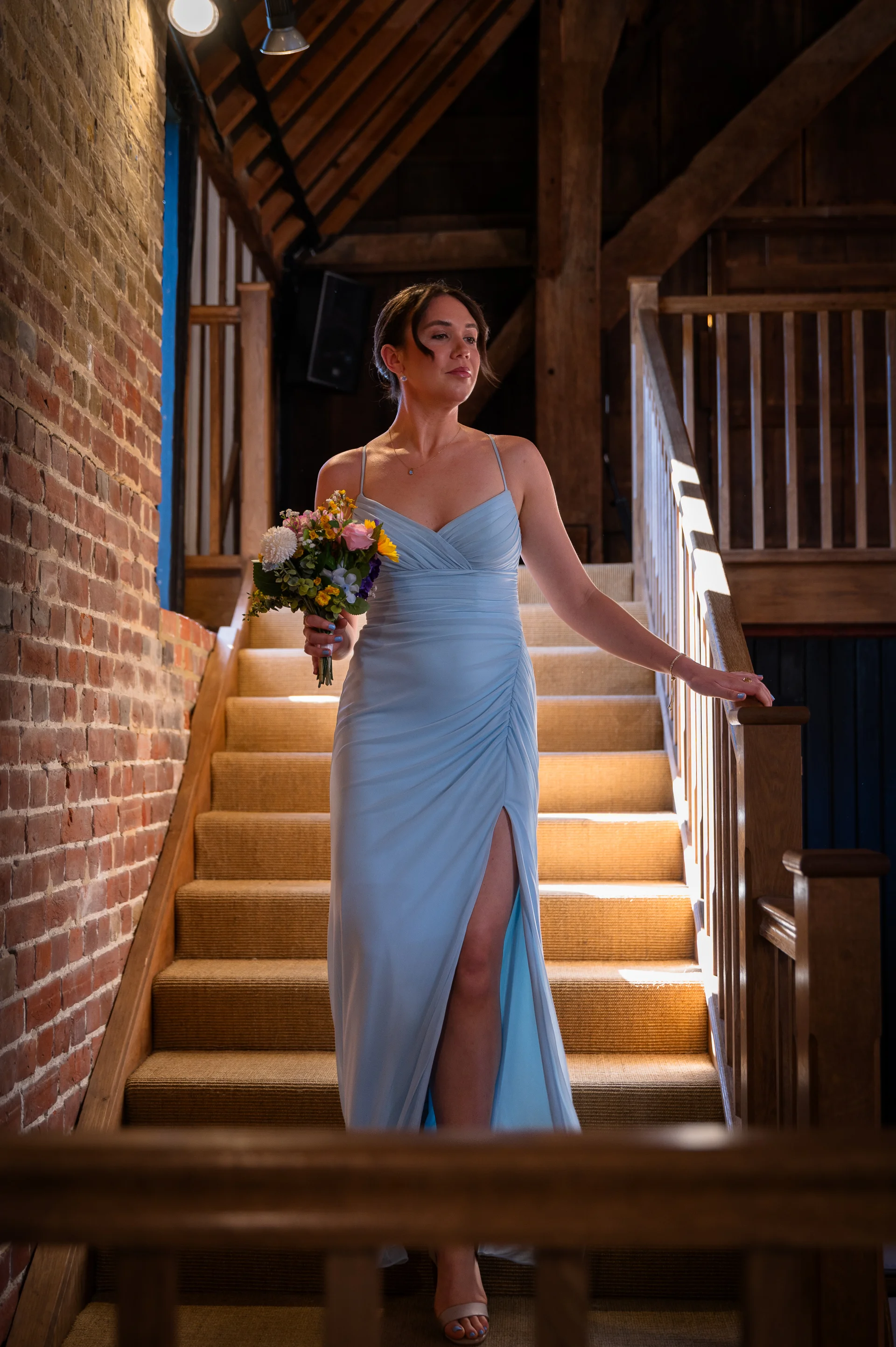 A bridesmaid in a dusty blue dress with a thigh split and ruched bodice descends a wooden staircase inside a rustic barn, holding a bouquet of colorful flowers.