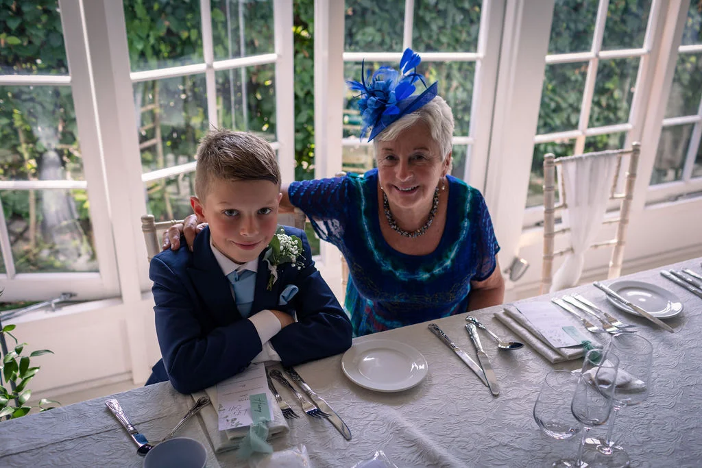 An elderly woman wearing a blue dress and fascinator sits next to a young boy in a blue suit at a wedding reception table.