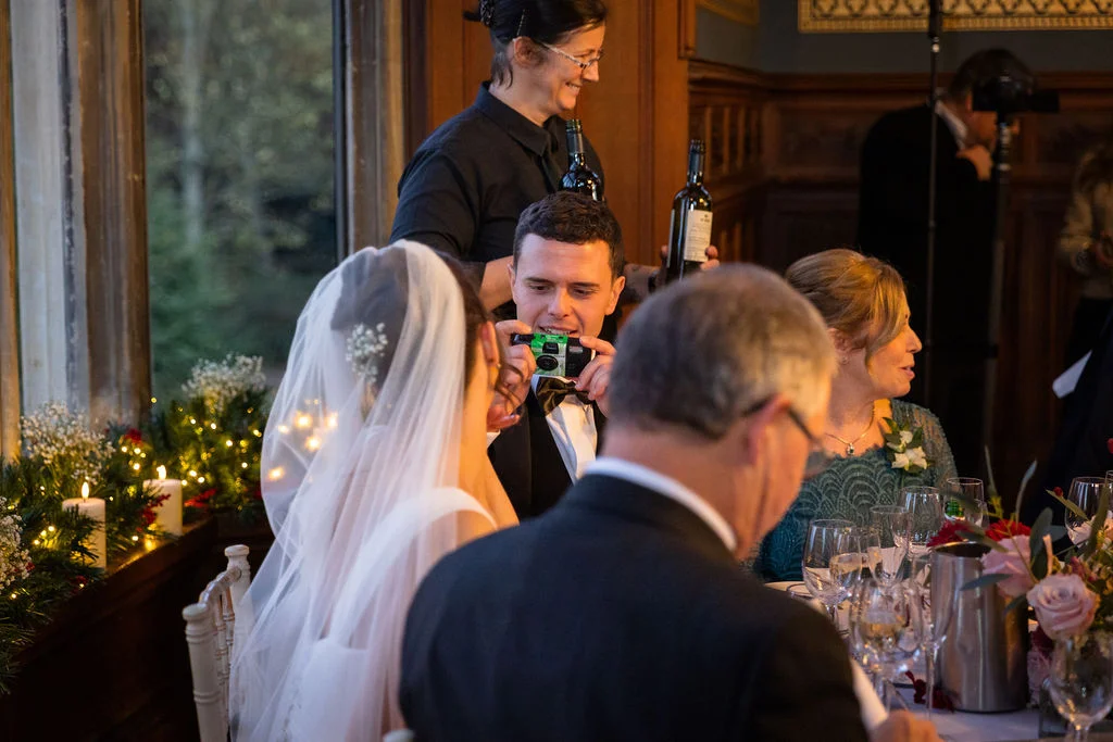 A groom smiles while taking a photo with a disposable camera at the top table during a candlelit wedding reception.