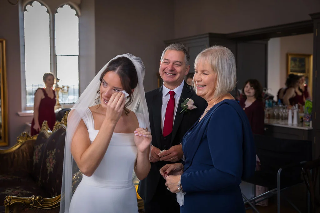 An emotional bride wipes away happy tears with a tissue while her parents stand proudly beside her in an elegant room during wedding preparations.