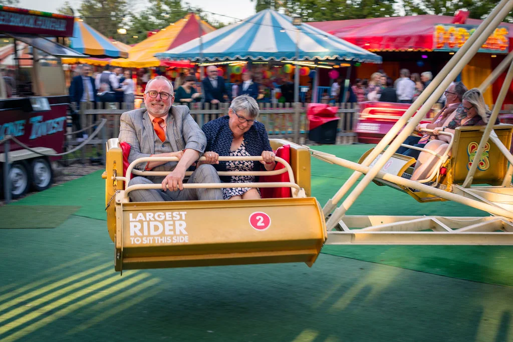 An older couple joyfully ride a vintage amusement park swing at a festival-themed wedding reception, surrounded by colourful fairground stalls.