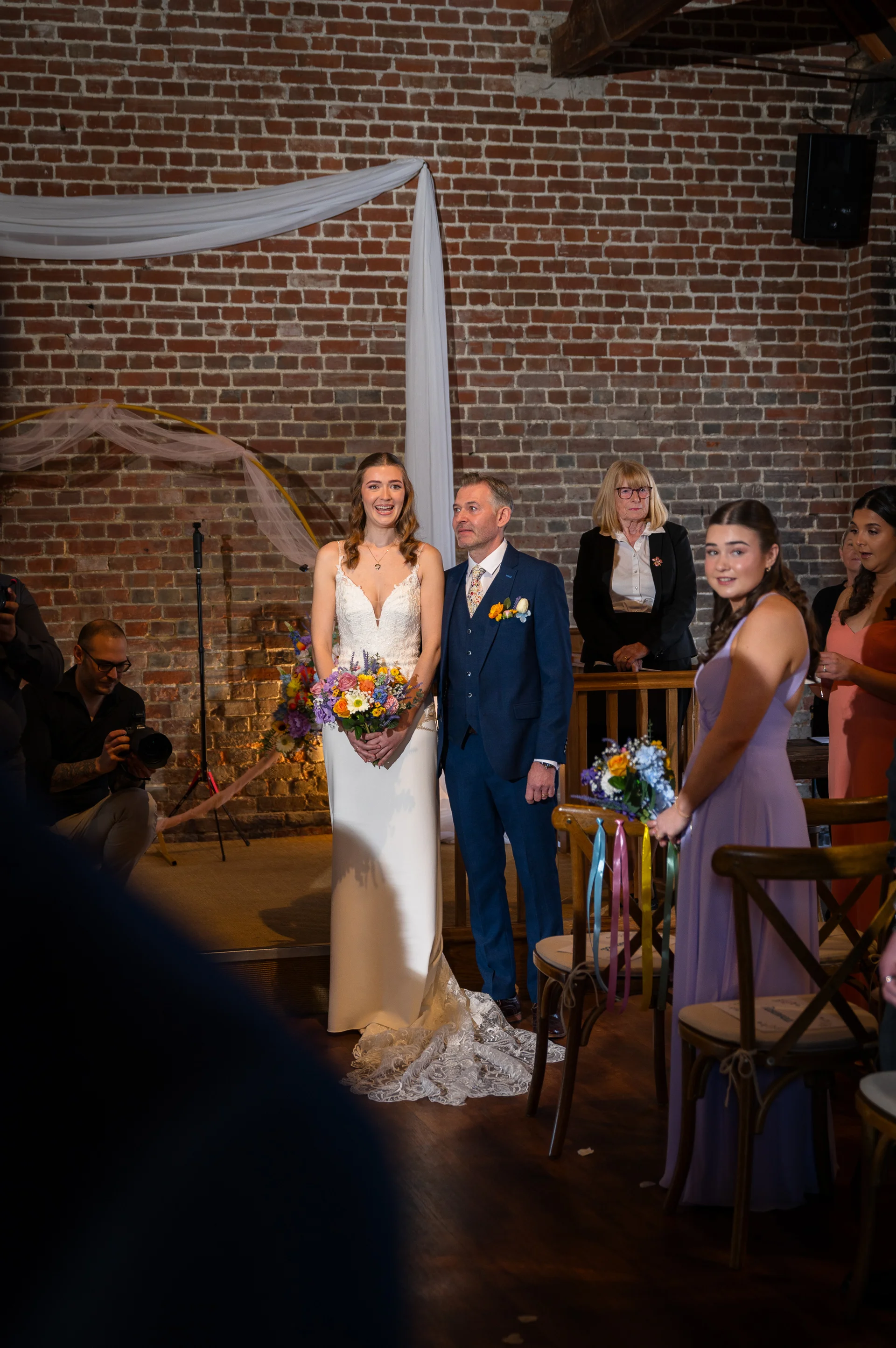 A joyful newly married couple stand together at the front of an exposed brick barn ceremony venue holding a colourful bouquet, with a bridesmaid watching closely from the aisle.