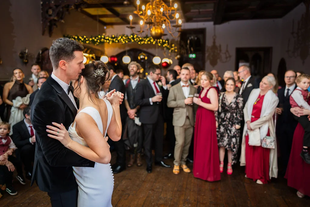 Newlyweds share their first dance in a grand hall with wooden floors and a glittering chandelier overhead while guests watch and mingle around the dance floor.
