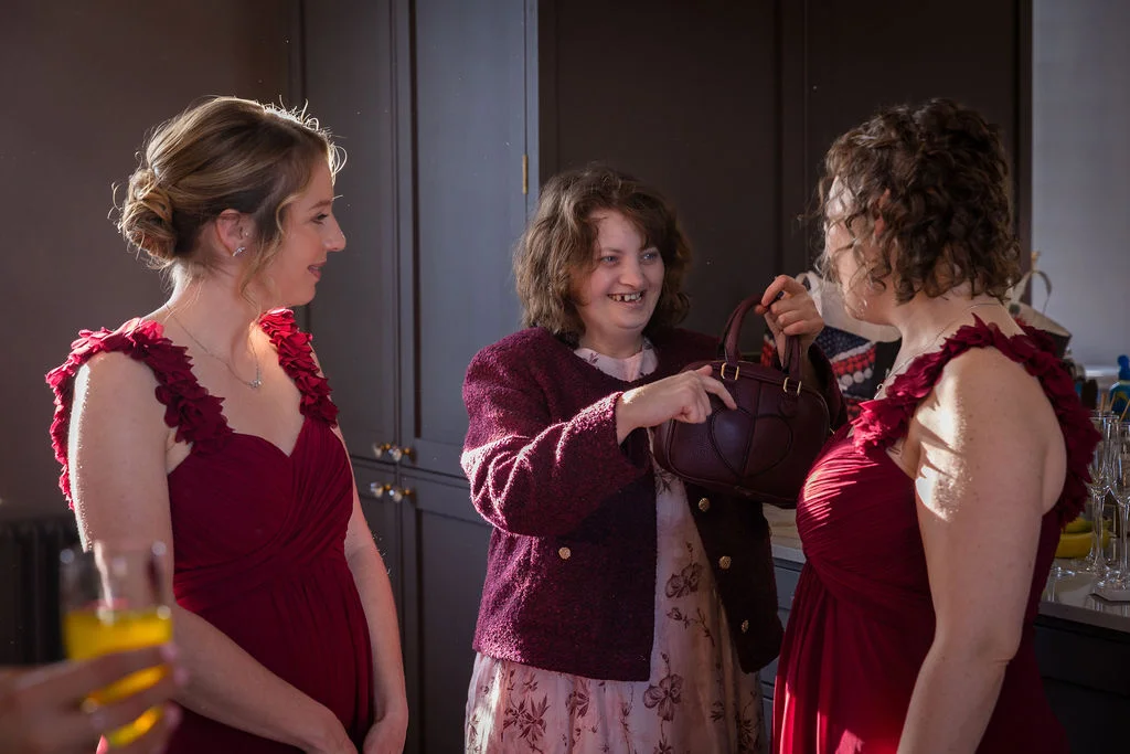 Three women in a room, with two bridesmaids wearing matching red dresses and another guest laughing while showing a small handbag during morning preparations.