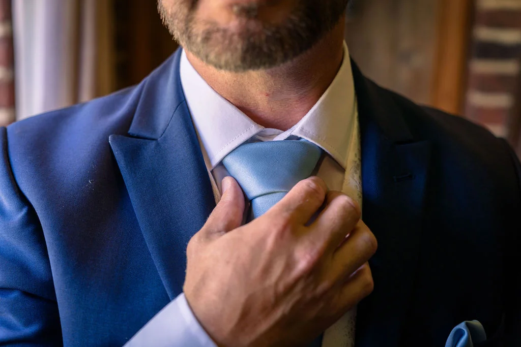 A groom in a blue suit carefully adjusts his blue tie during morning preparations on his wedding day.