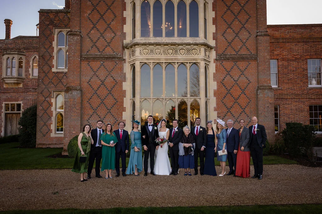 A large wedding party and family members pose for a formal group photograph outdoors on a gravel path in front of a grand red brick manor house during golden hour.
