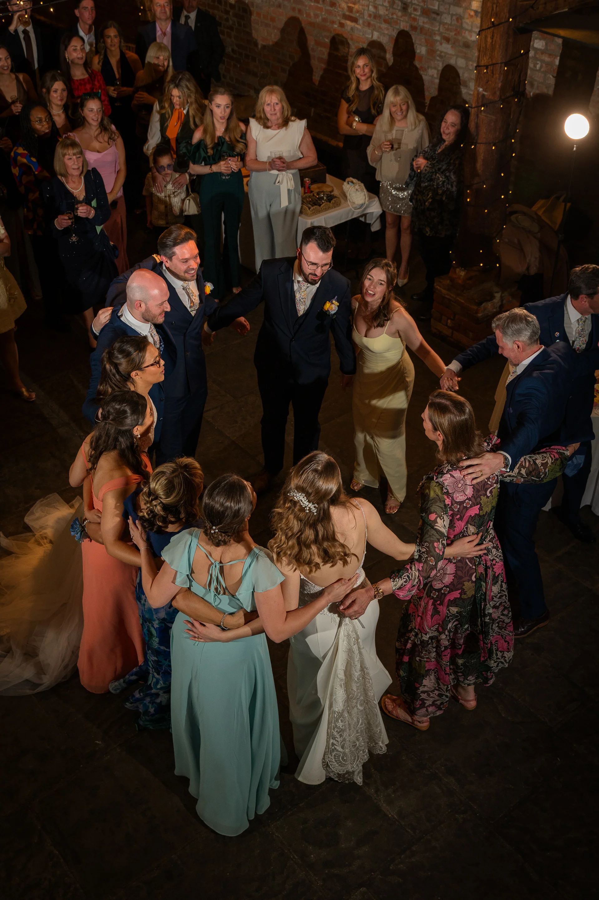 A high-angle view shows the newlyweds and their wedding guests holding hands and dancing in a circle inside a rustic brick barn lit by festoon lights.