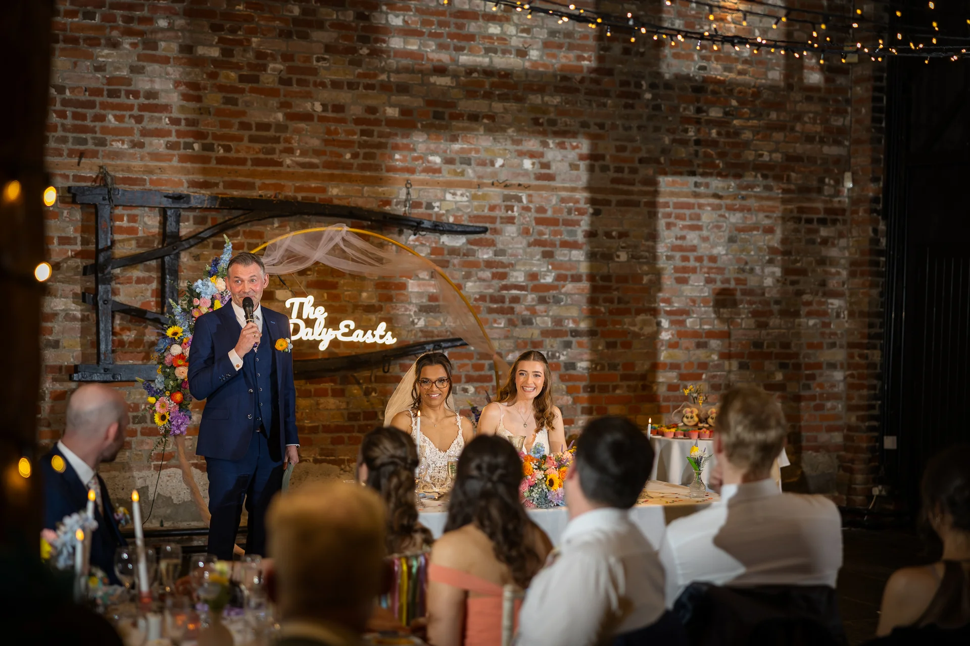 A guest delivers a speech with a microphone at the top table in an exposed brick barn venue, while the two smiling brides look on seated below a custom neon sign.