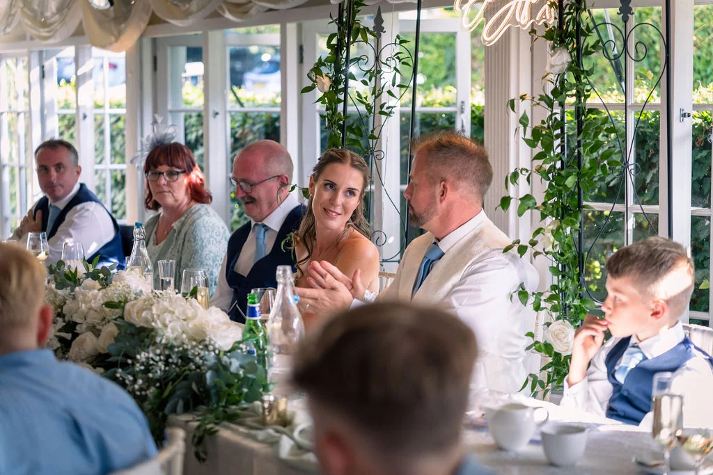 The bride and groom share a tender glance while seated at the top table during a sunlit wedding breakfast in a conservatory.