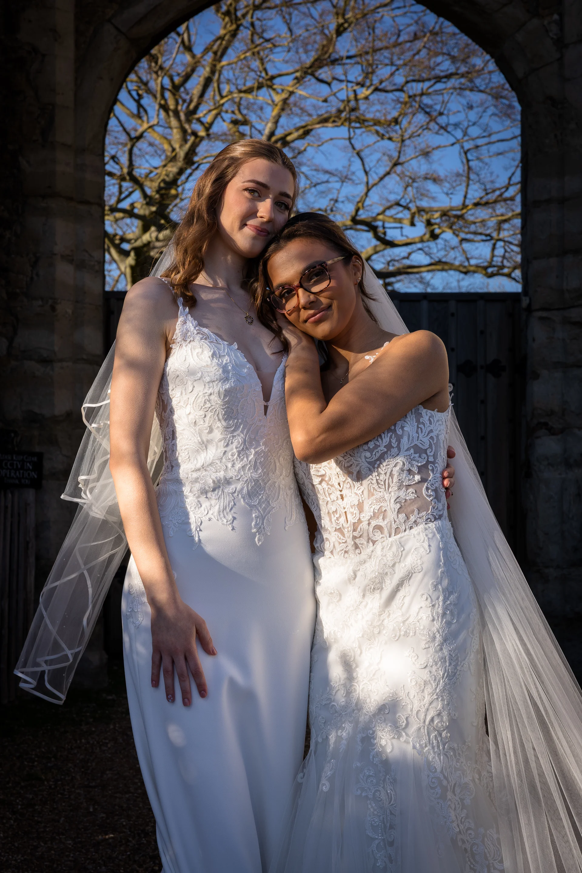 Two smiling brides in white lace wedding gowns stand closely together, framed by a historic stone archway under a bright blue sky.