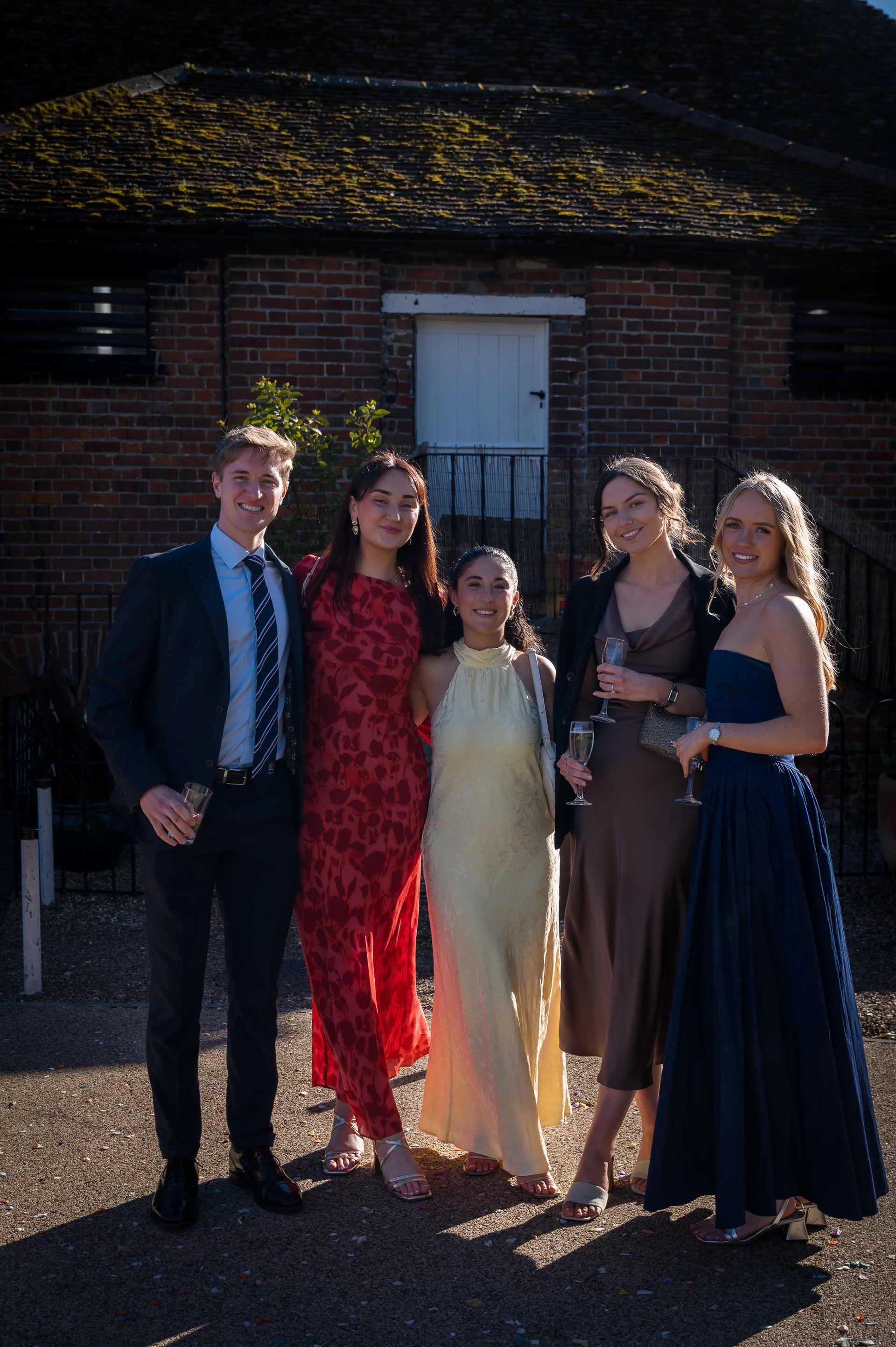 A group of five wedding guests pose with drinks outside against a rustic brick barn exterior on a sunny afternoon.