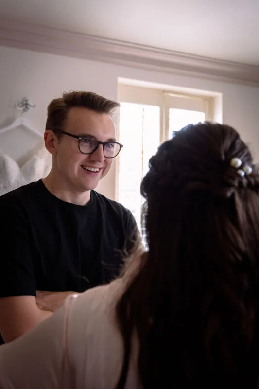 A smiling man wearing glasses and a black t-shirt looks warmly at a partner with dark wavy hair and pearl hair pins during morning preparations.