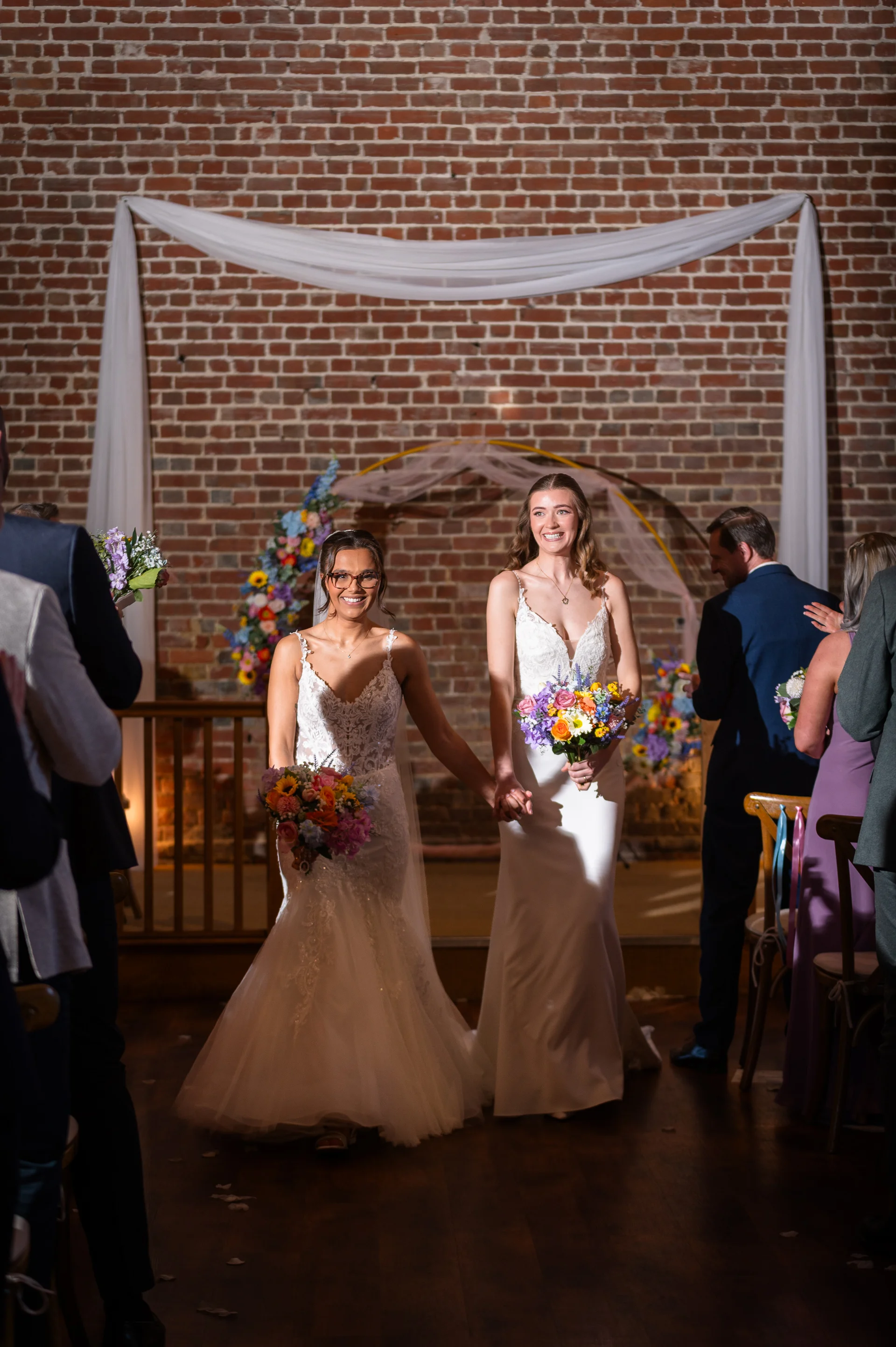 Two smiling brides holding hands and walking down the aisle together after their ceremony in an exposed brick barn venue.