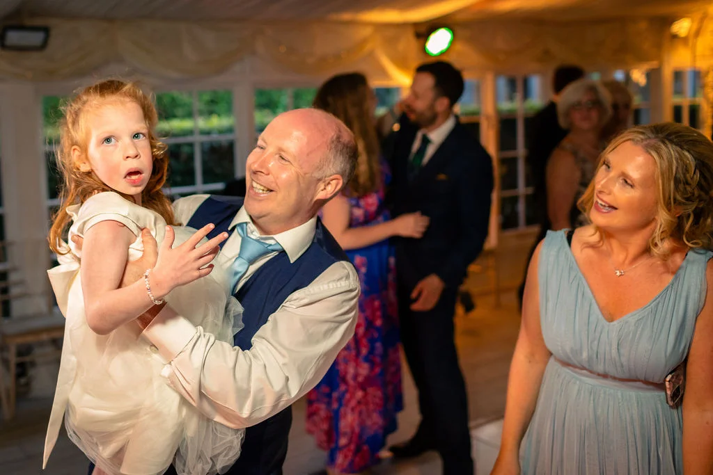 A smiling guest holds a young girl in a white dress on the dance floor during an evening wedding reception inside a marquee.