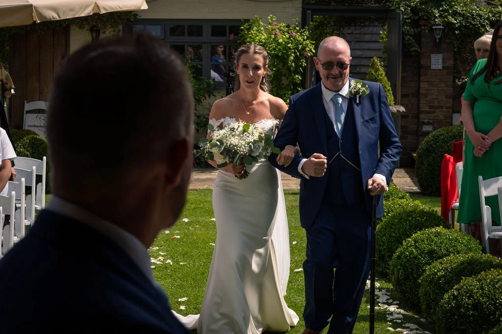 The bride walks down an outdoor garden aisle holding a white floral bouquet, linked arm-in-arm with her father as the groom looks on from the foreground.