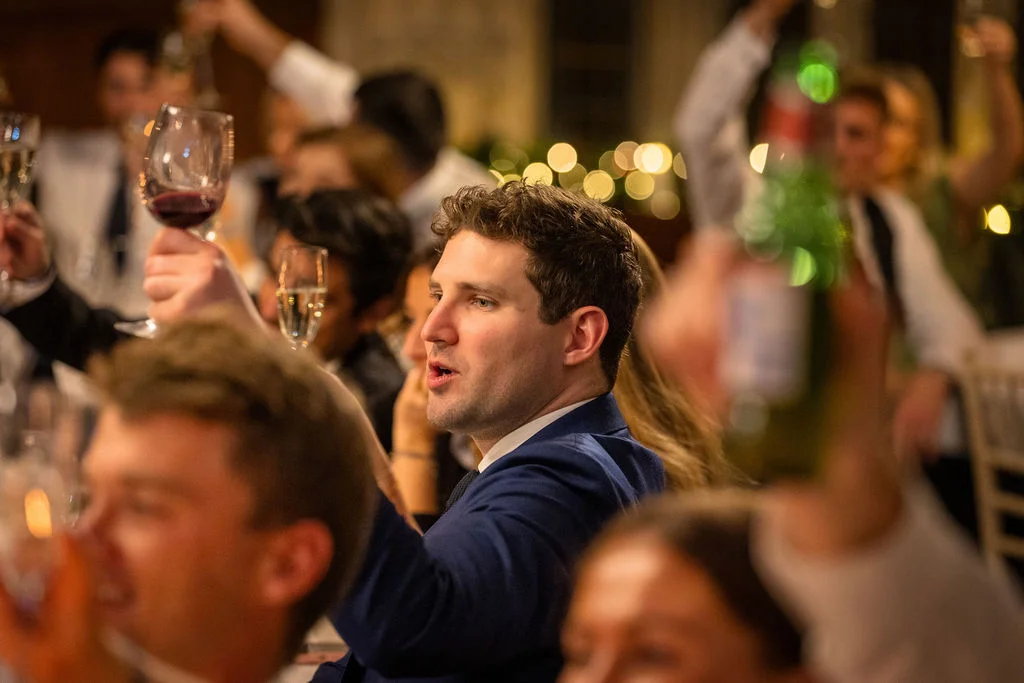 A joyful wedding guest in a blue suit raises his glass to toast during the wedding reception speeches, surrounded by cheering guests and warm candlelit ambiance.