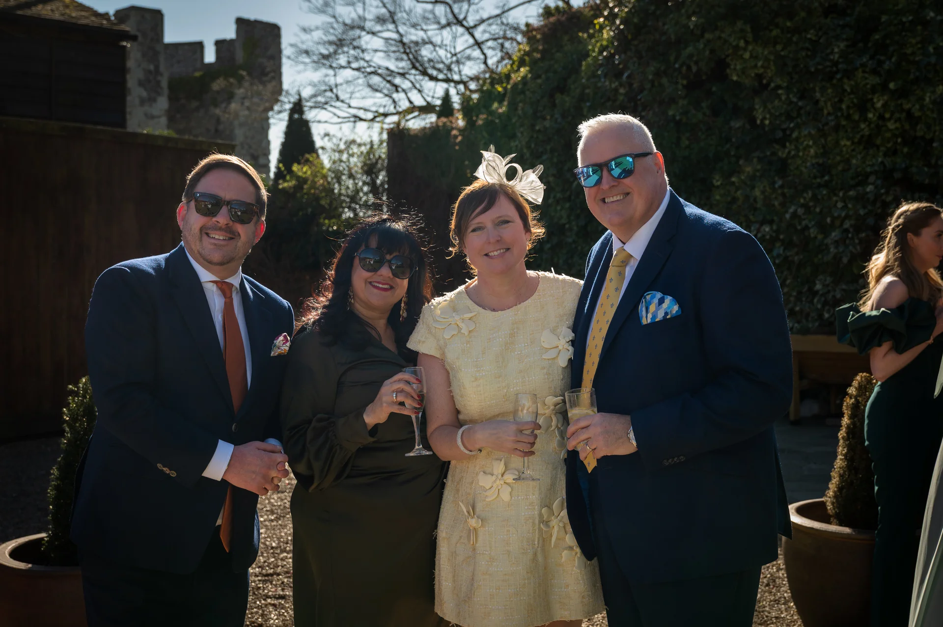 A group of smartly dressed wedding guests wearing sunglasses and holding drinks enjoy the sunshine outdoors during a reception at a historic venue.