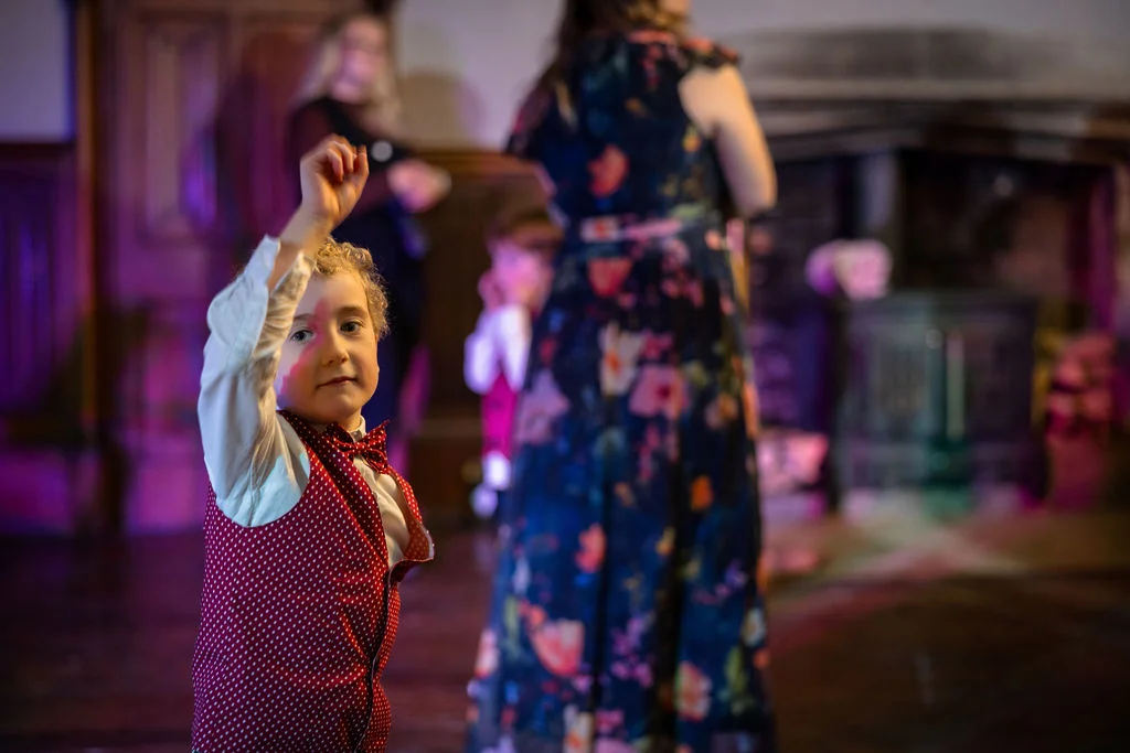 A young page boy wearing a red polka dot waistcoat and bow tie raises his hand while dancing at an evening wedding reception.