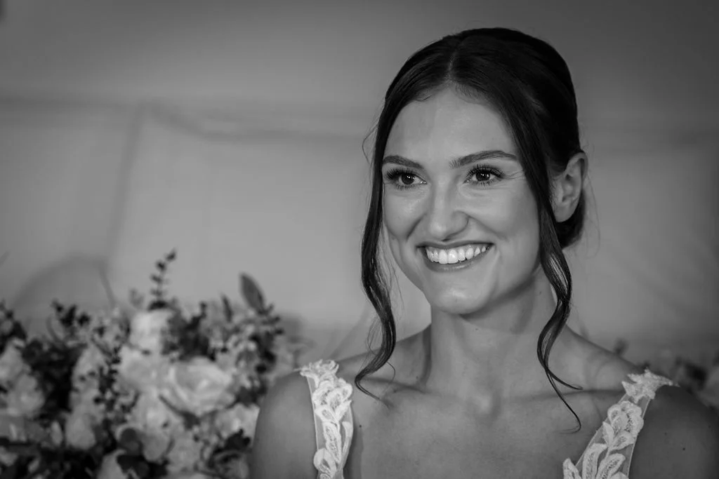 A close-up black and white portrait of a smiling bride wearing a lace dress with floral applique and her hair styled in an updo with face framing tendrils.