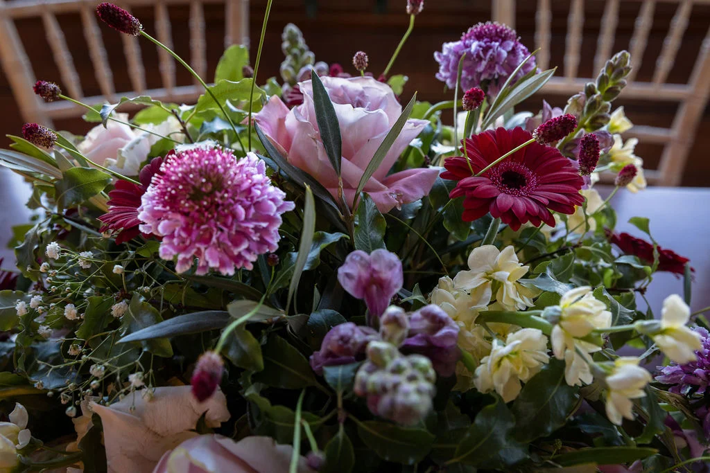 A close up view of a rich floral arrangement featuring deep plum, pink, and burgundy blooms mixed with delicate white flowers and green foliage.