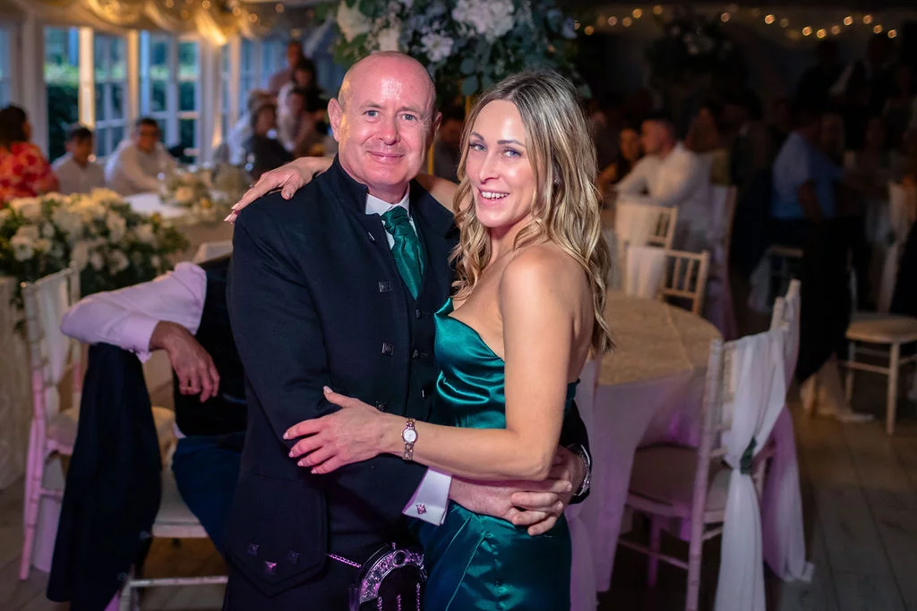 A smiling couple embraces during an evening wedding reception, with the groom wearing a traditional kilt and green tie while the partner wears an emerald green satin dress.