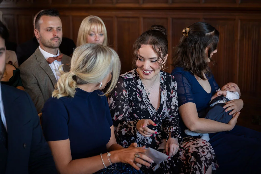 Wedding guests smiling and chatting warmly inside a wood-panelled room, with a baby nestled safely in a family member's arms.