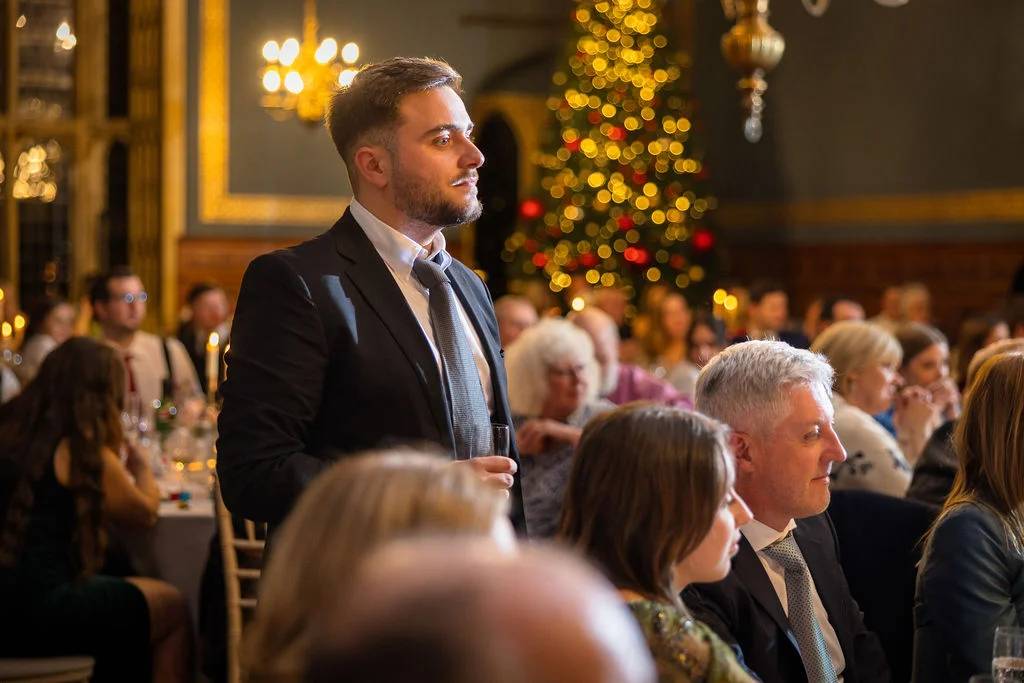 A man in a black suit delivers a speech during a festive winter wedding reception, with a glowing Christmas tree in the background.