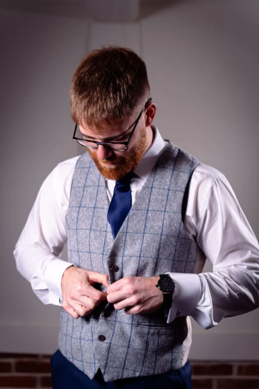 A groom with a ginger beard wearing a checked grey waistcoat and a dark blue tie fastens his buttons while getting ready.