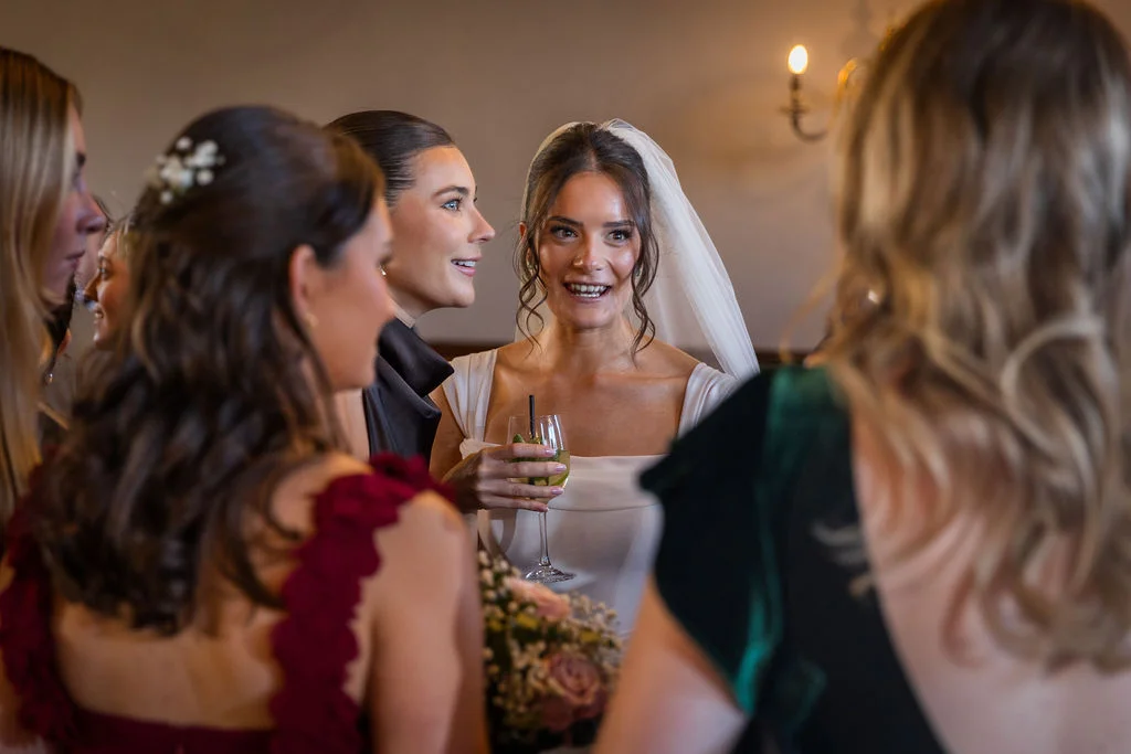 A smiling bride wearing a veil and holding a glass and bouquet laughs while chatting with her bridesmaids at the drinks reception.
