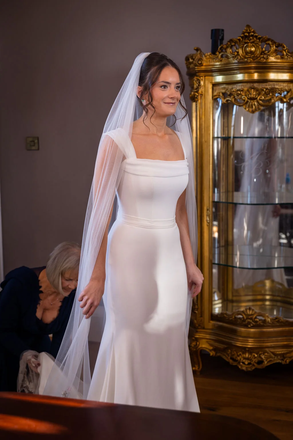 A smiling bride wearing a crepe gown with a square neckline and a long veil stands proudly while someone adjusts her dress indoors.
