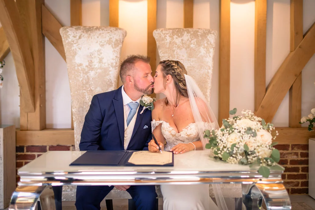 Newlyweds share a romantic kiss while seated at a glossy white table during the signing of their marriage certificate inside an oak-framed barn.