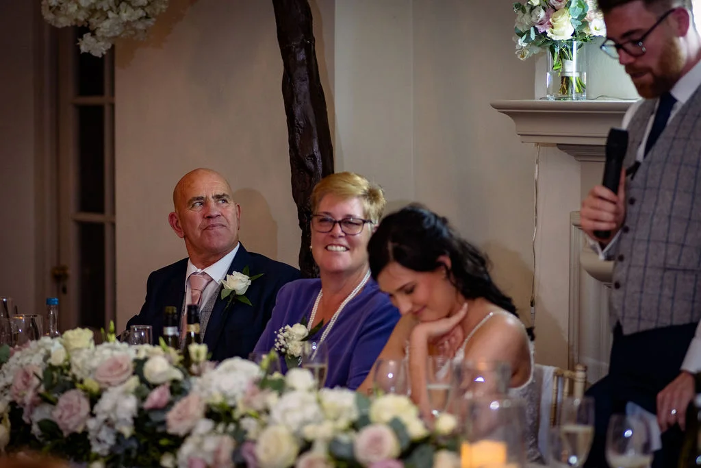 Family members and a bride seated at the top table smile and laugh during the wedding speeches while a guest speaks into a microphone.