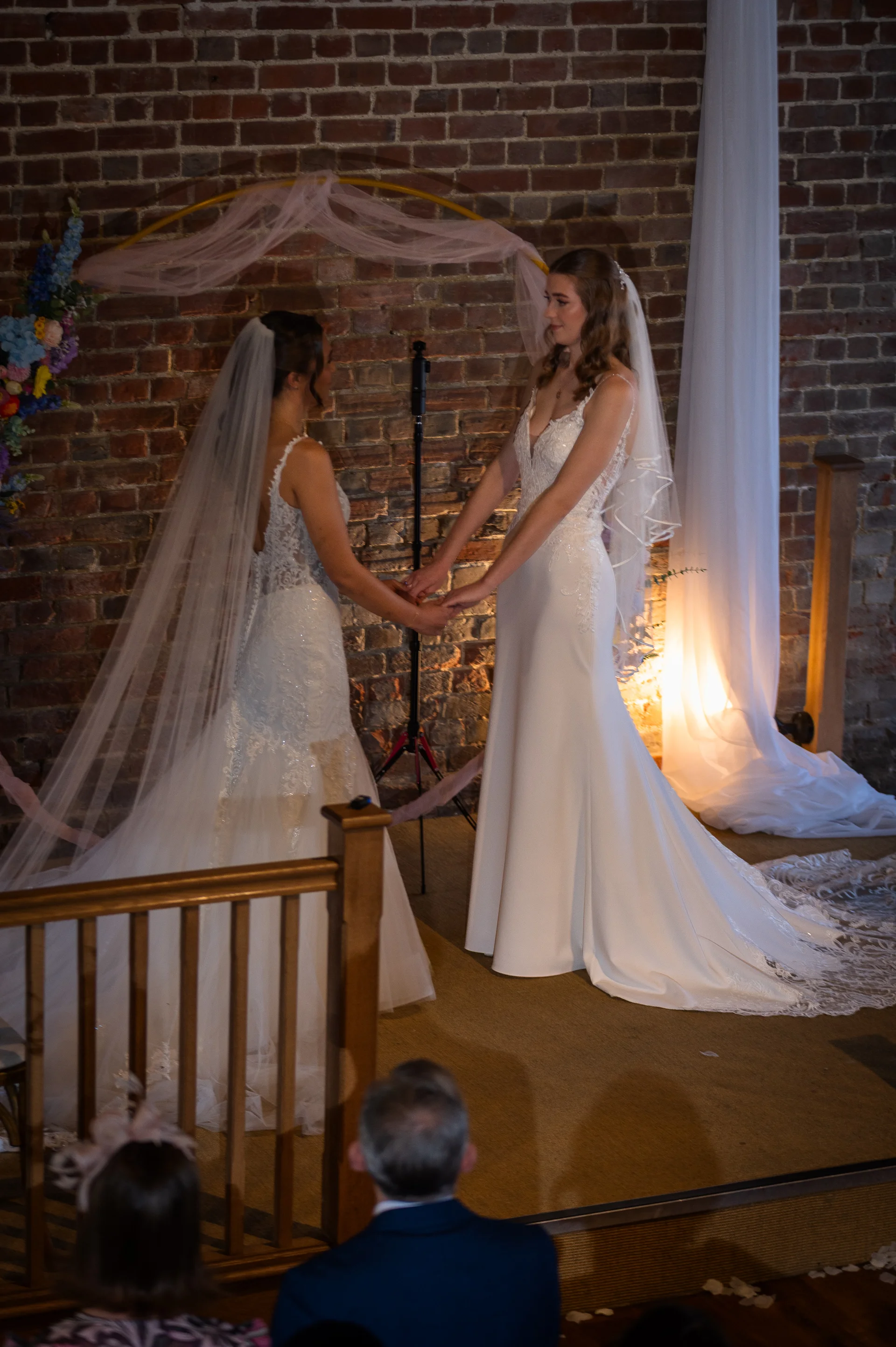 Two brides holding hands during their ceremony in front of an exposed brick wall inside a rustic barn venue.