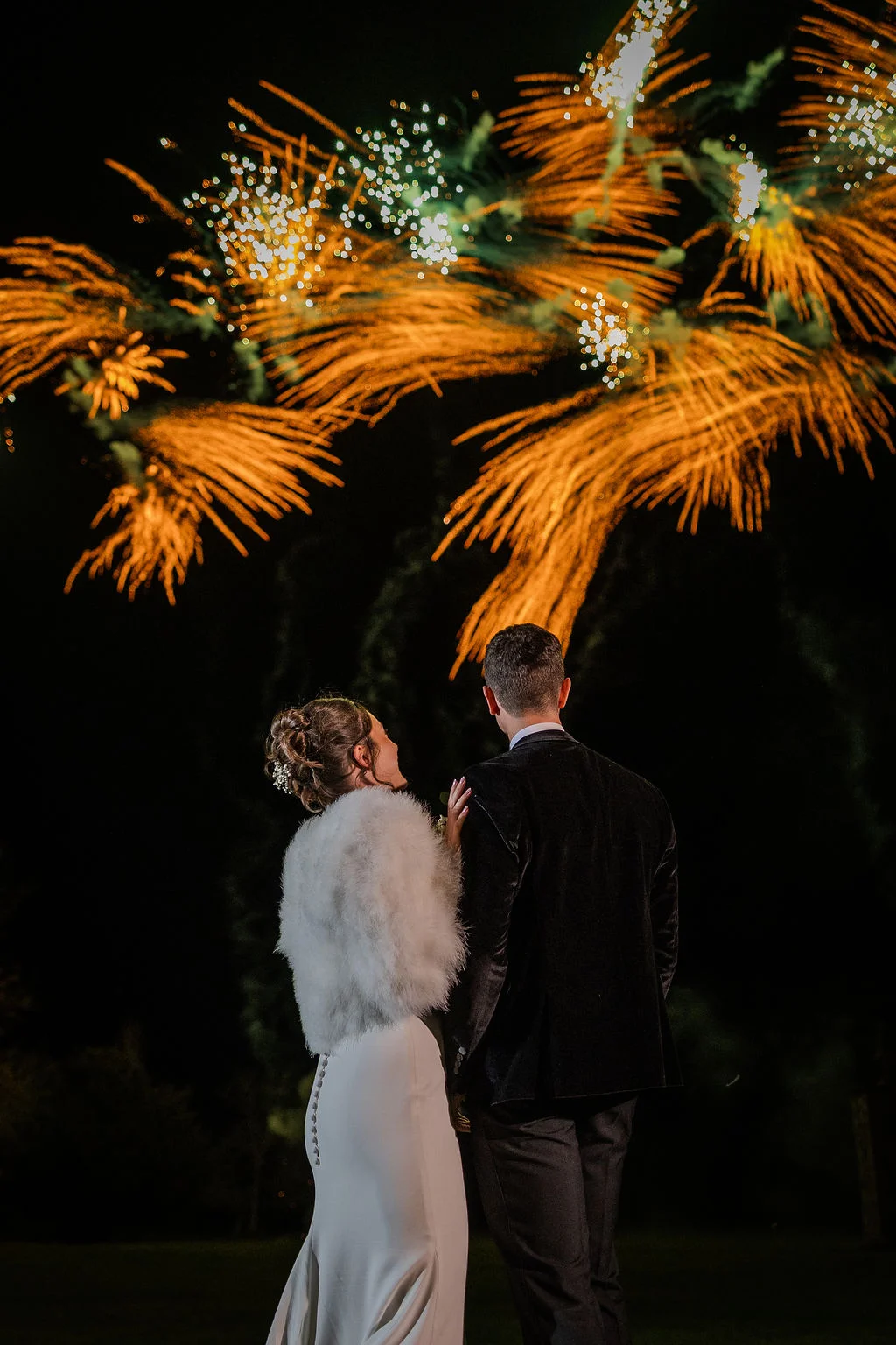 Newlyweds stand back to back against the dark night sky, watching a vibrant golden fireworks display light up overhead.