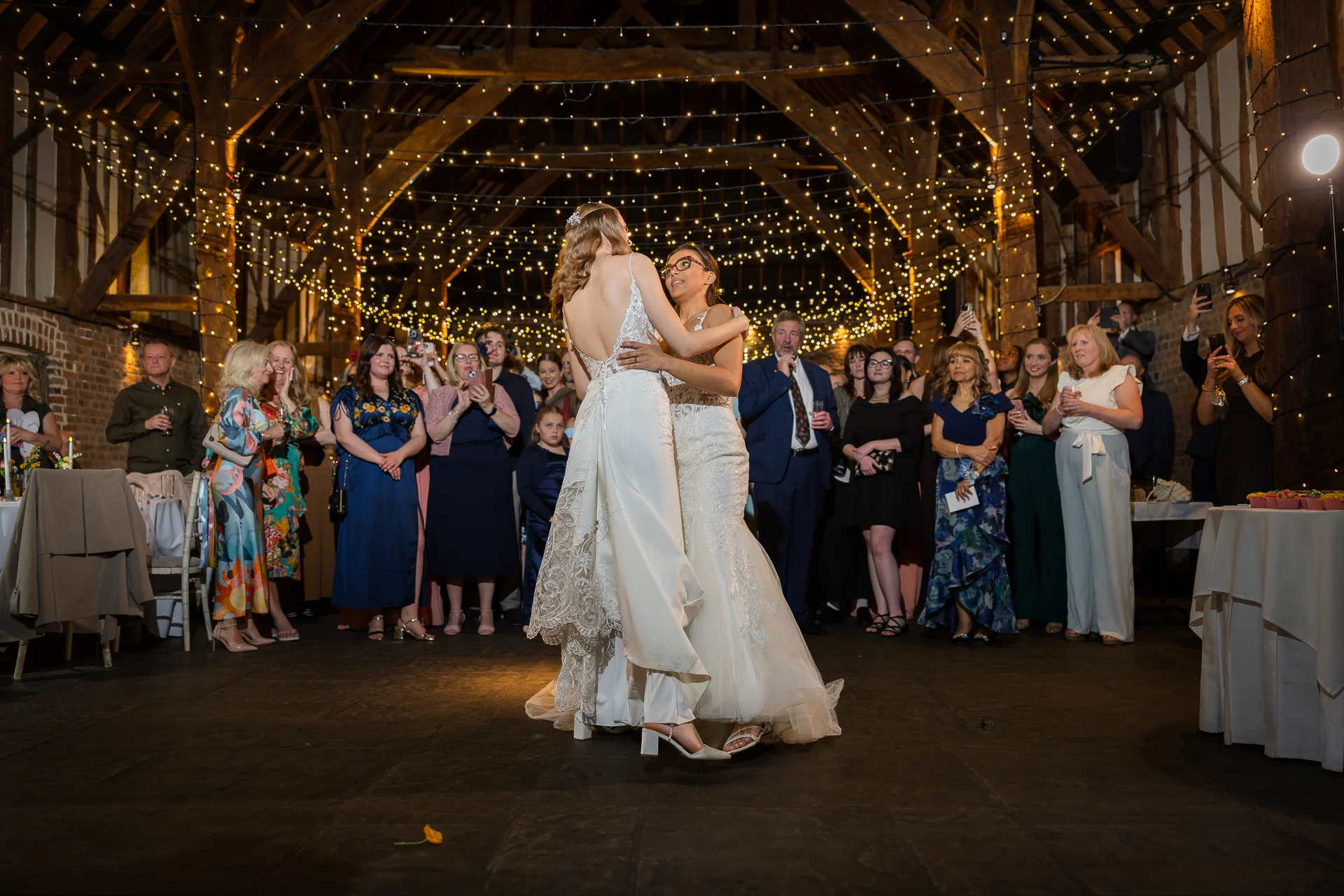 Two brides share their first dance in the centre of a rustic, oak-framed barn illuminated by a vast canopy of fairy lights as guests watch on.