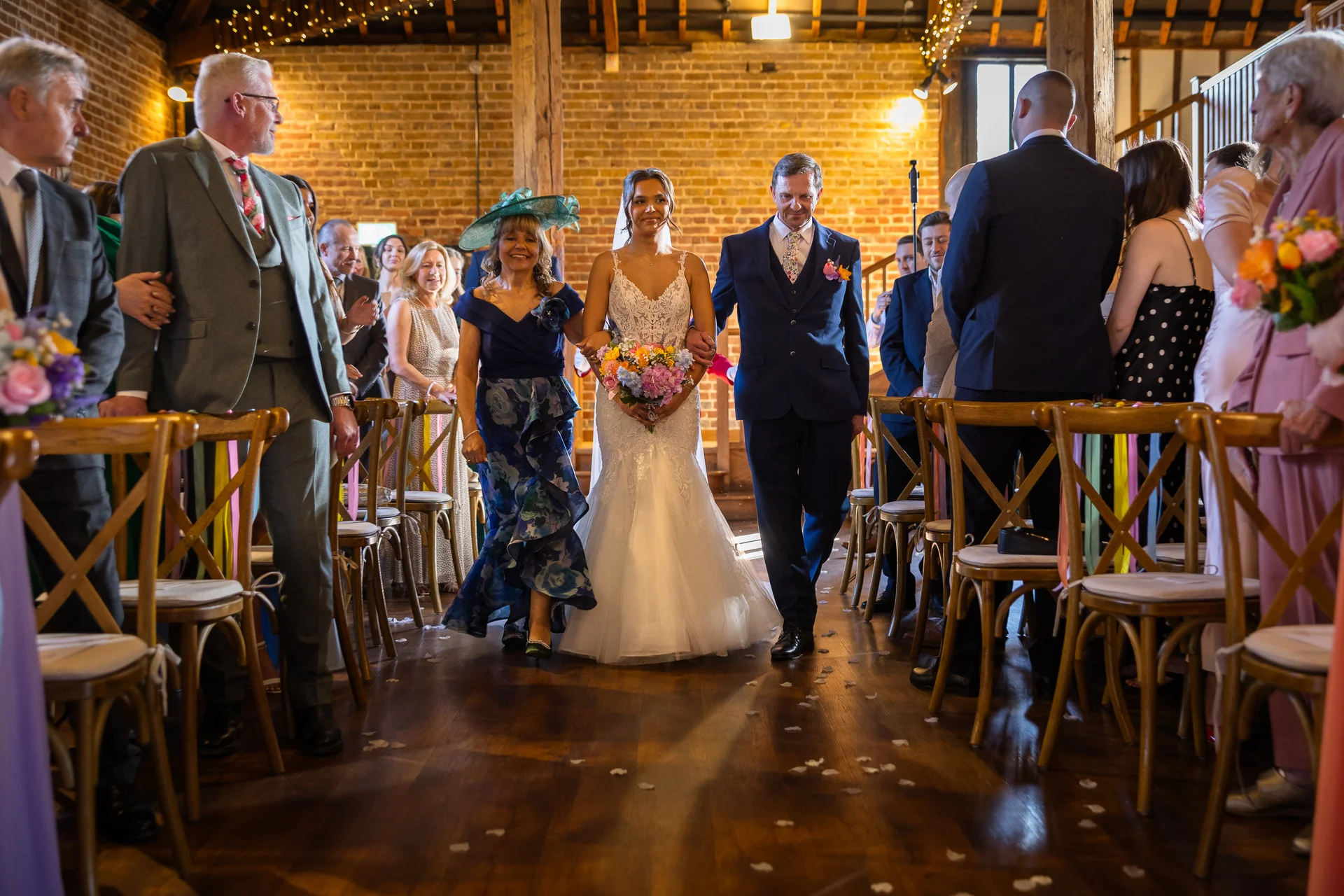 The bride walks down the aisle in a rustic brick barn accompanied by her parents, holding a colourful bouquet while guests look on from wooden chairs.