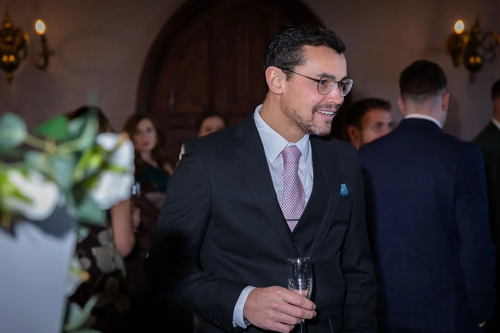 A smiling groom wearing a black suit, white shirt, and patterned tie holds a champagne glass while mingling with guests during a wedding reception.