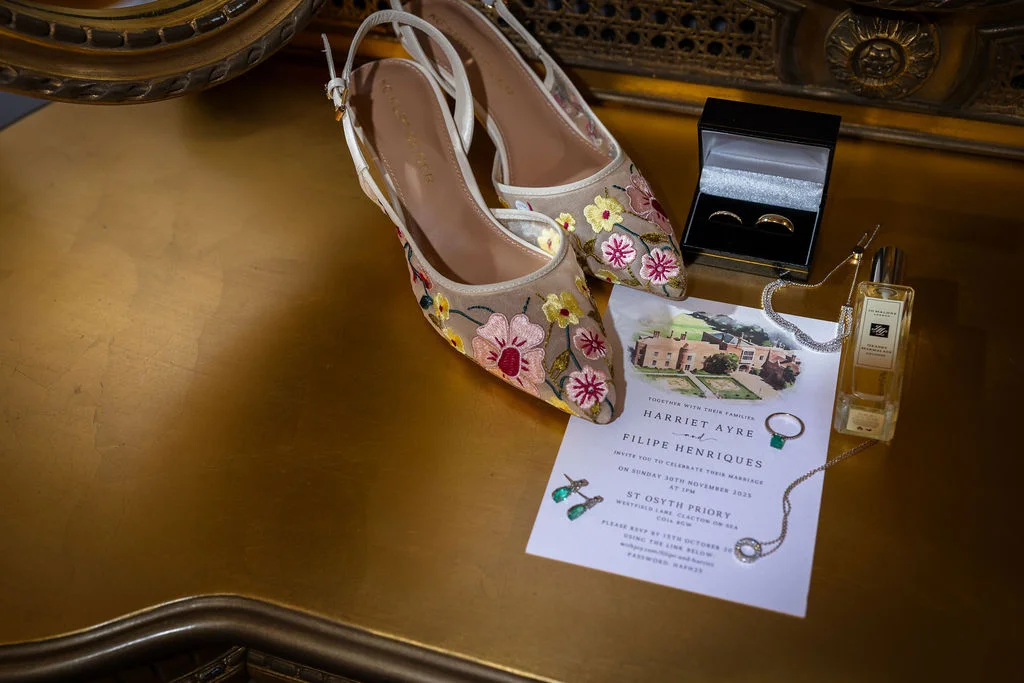 An elegant bridal flat lay on a gold surface featuring floral embroidered slingback heels, an illustrated wedding invitation, wedding rings in a black box, jewellery, and a bottle of perfume.