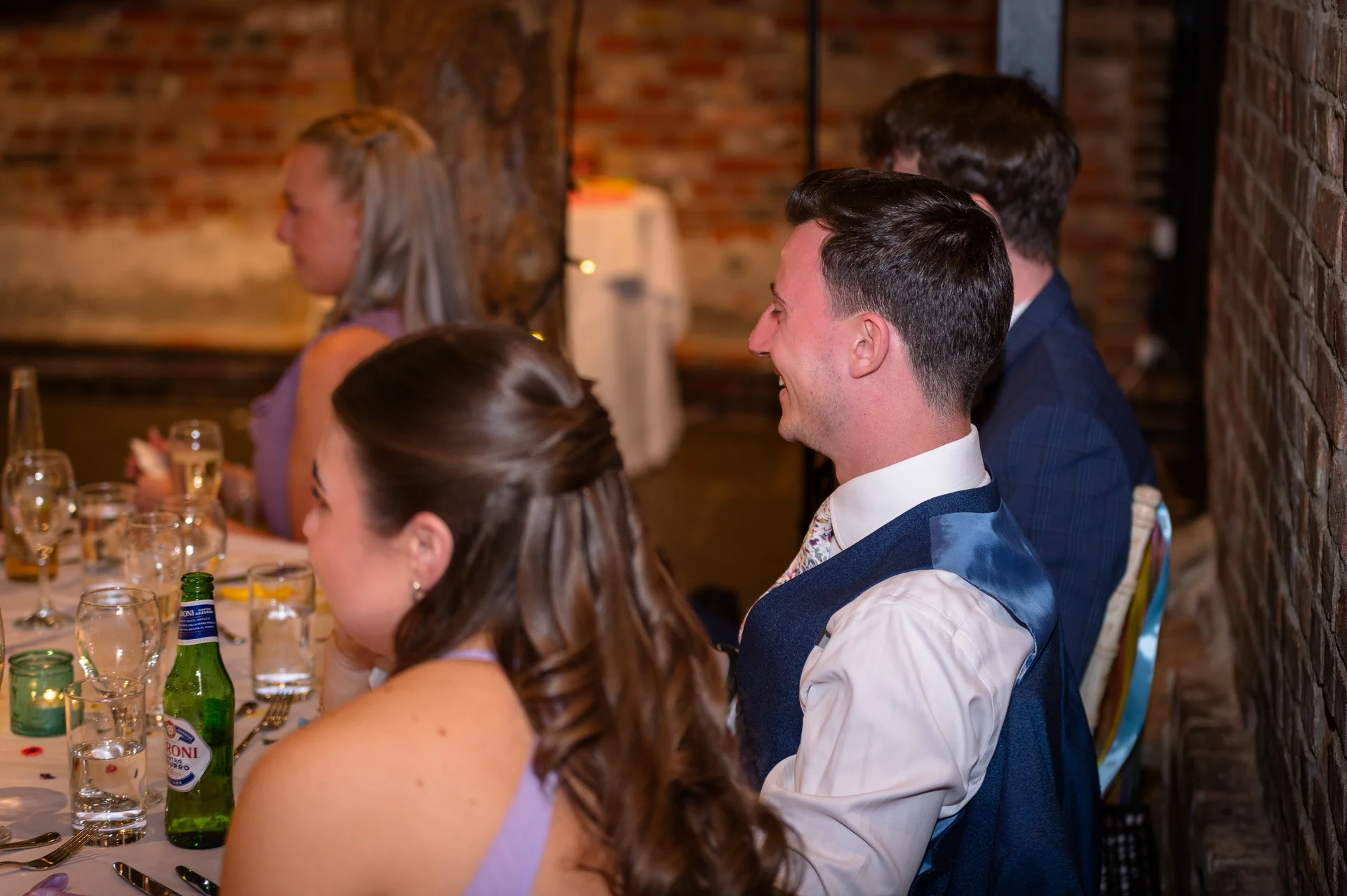 Wedding guests seated at a banquet table indoors with exposed brick walls laugh heartily during the wedding speeches.